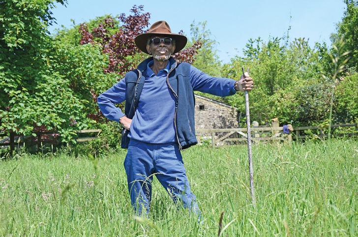 A black man leaning on a wooden staff in a grass meadow