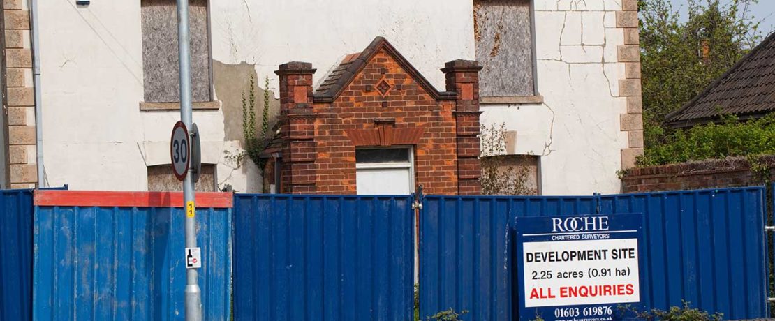 A run-down house behind bright blue hoardings with a sign for development