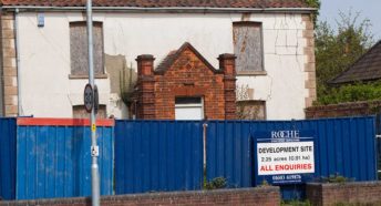 A run-down house behind bright blue hoardings with a sign for development