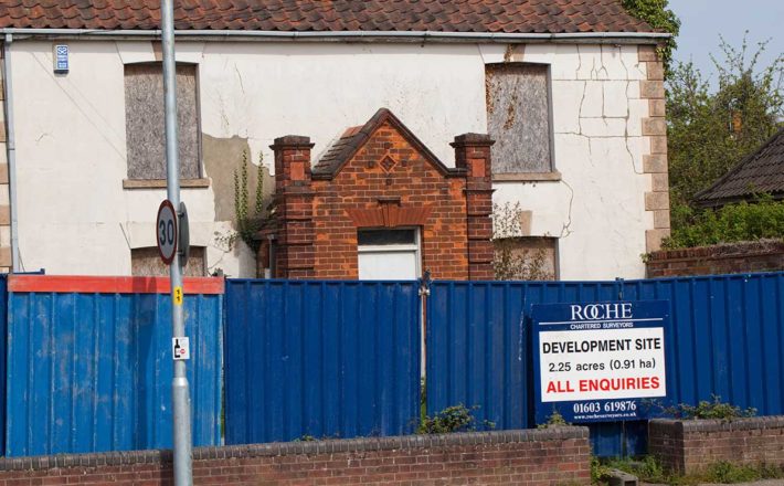 A run-down house behind bright blue hoardings with a sign for development