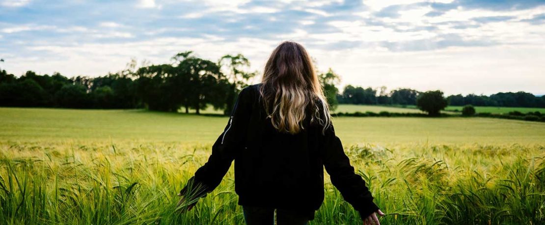 A teenager stands at the edge of a field with her back to us