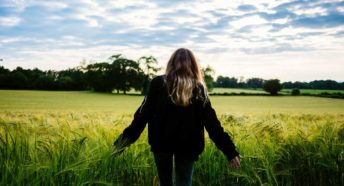 A teenager stands at the edge of a field with her back to us