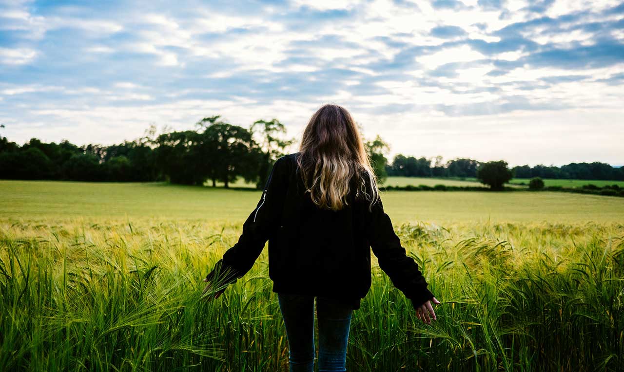 A teenager stands at the edge of a field with her back to us