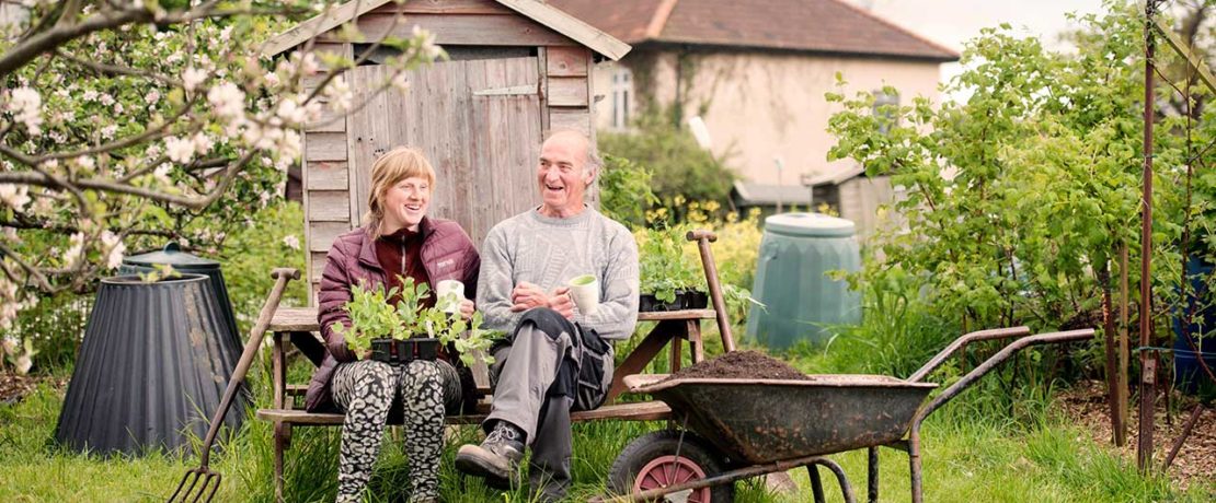 A man and a woman sit on a bench holding plants and tea mugs in a flourishing allotment