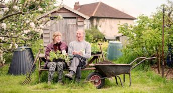 A man and a woman sit on a bench holding plants and tea mugs in a flourishing allotment
