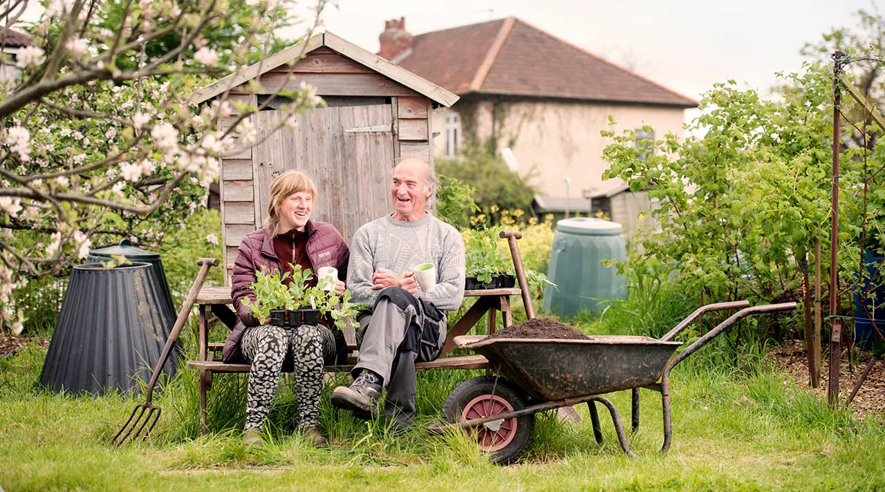 A man and a woman sit on a bench holding plants and tea mugs in a flourishing allotment