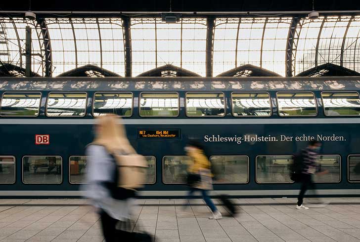 People rush in front of a train with German text on the side in a dramatic station