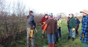 A group of people with hedgelaying equipment standing next to a hedge in winter