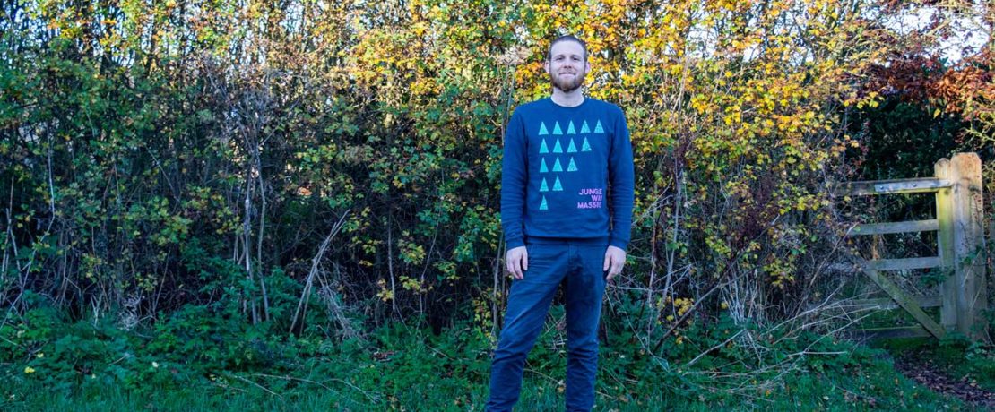 A white man in a blue jumper stands in front of a bush with bright autumn leaves