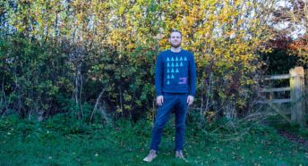 A white man in a blue jumper stands in front of a bush with bright autumn leaves