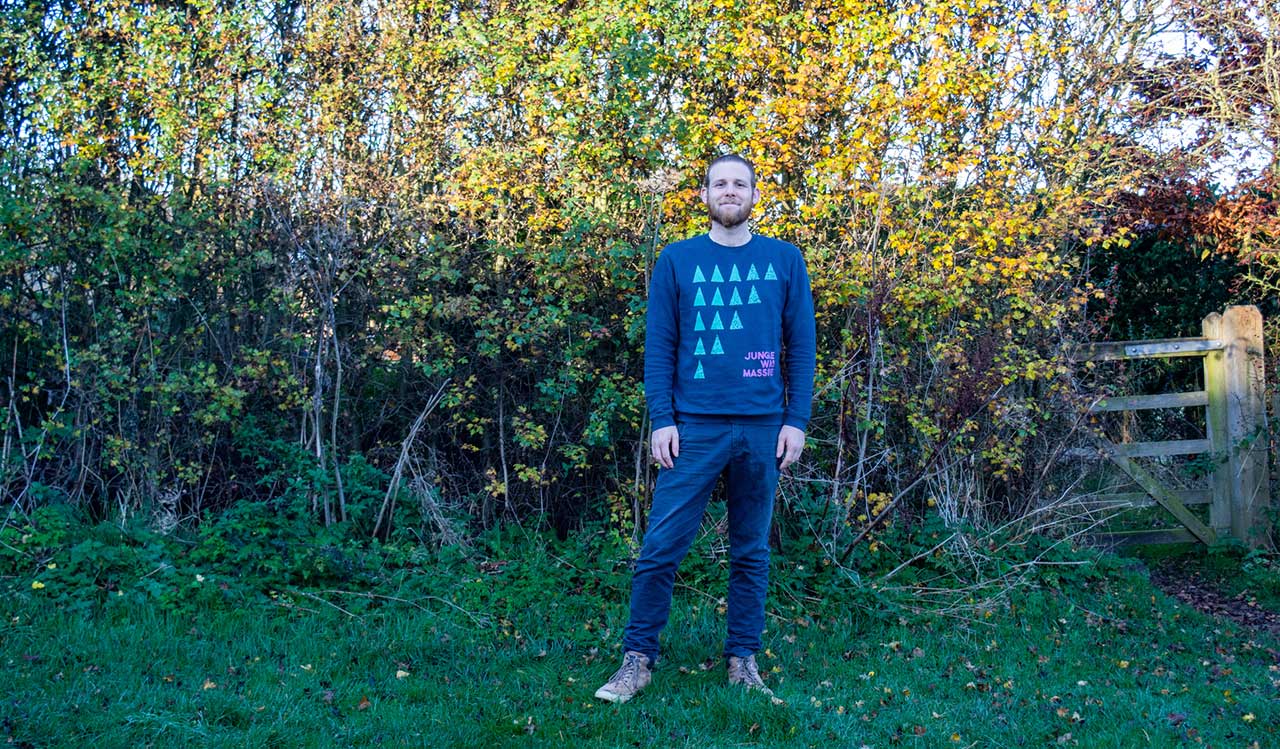 A white man in a blue jumper stands in front of a bush with bright autumn leaves