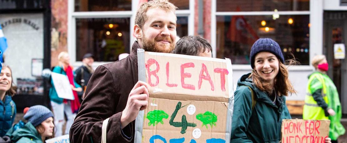 Two young people in warm clothes walk along a busy street with placards; one says Bleat for Peat