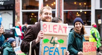 Two young people in warm clothes walk along a busy street with placards; one says Bleat for Peat