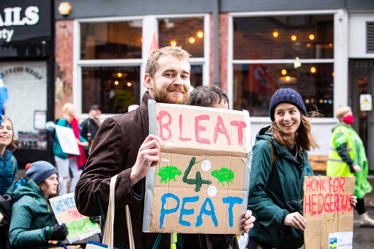 Two young people in warm clothes walk along a busy street with placards; one says Bleat for Peat