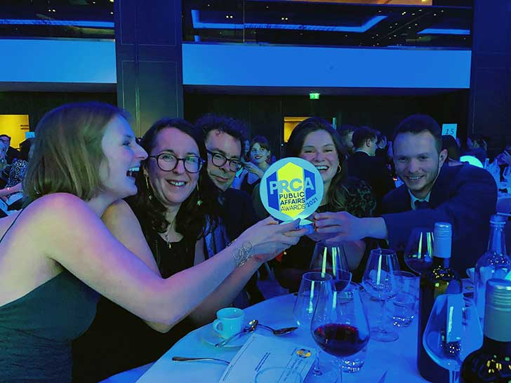 Five people lean around a round table to share holding a bright award that says PRCA