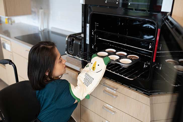A lady in a wheelchair leans forward, wearing an oven glove, to push cakes into the oven