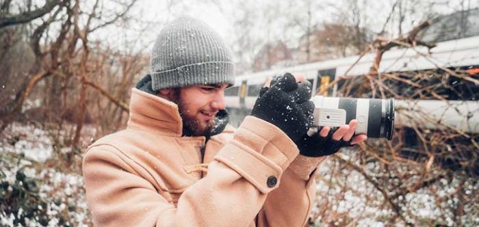 A man in a warm duffel coat holds a large camera and snaps a train