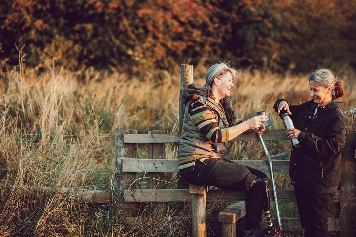 Two middle aged woman, one with prosthetic leg, stopping by stile for a drink from a flask