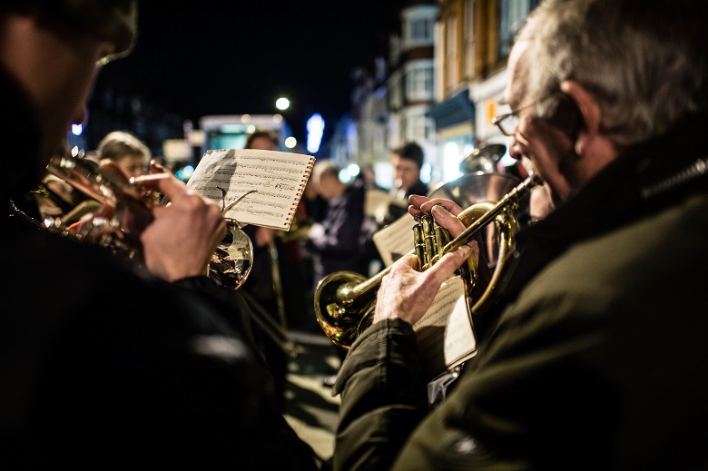 Looking over the shoulder of a brass band playing Christmas music carols, outdoors at night