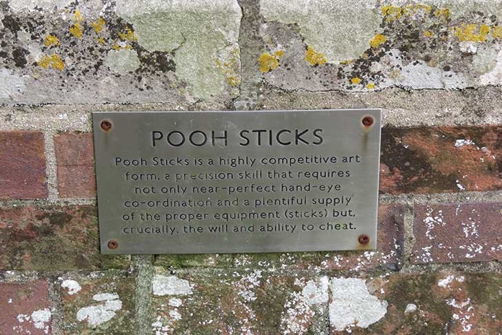 A metal plaque attached to a brick wall, with text about the craft of a game of Pooh Sticks