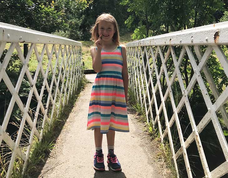 A young girl in a sundress holds a small stick and stands on a bridge