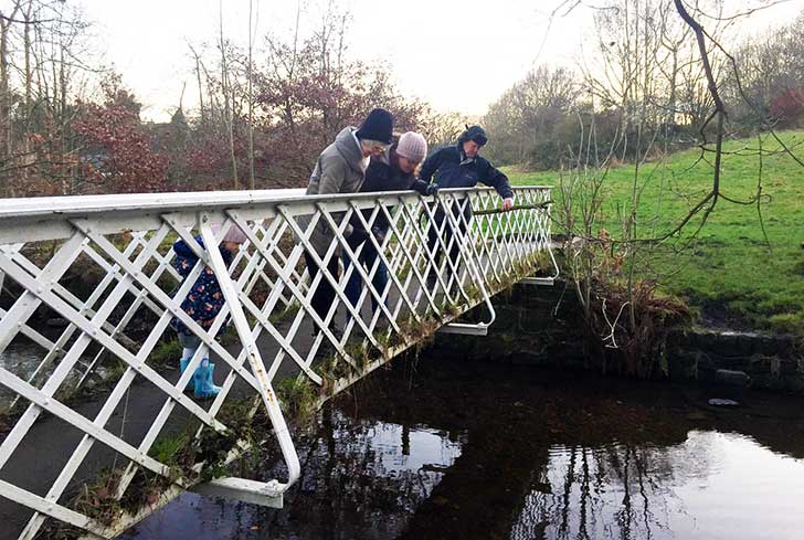 A senior couple and a younger woman leaning over a white metal bridge looking down at a stream