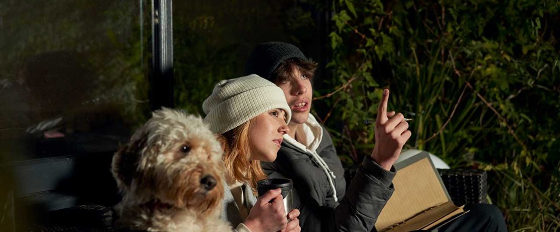 A young man, woman and a dog sit side by side in a dark garden and look upwards