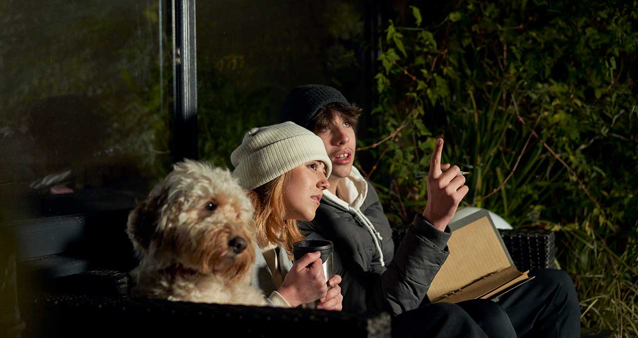 A young man, woman and a dog sit side by side in a dark garden and look upwards at the stars