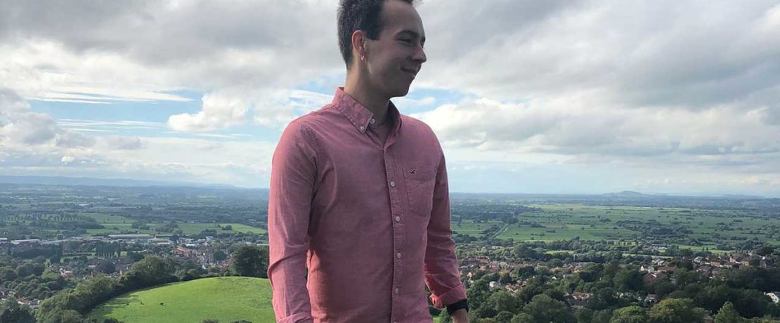 A person in a pink shirt and jeans with a dramatic green landscape behind