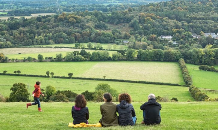 Three women and man watch young boy running on hill with view of fields and trees beyond.