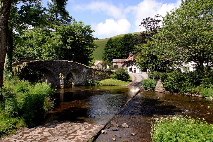 Old stone bridge by river ford on sunny day with white cottage surrounded by trees