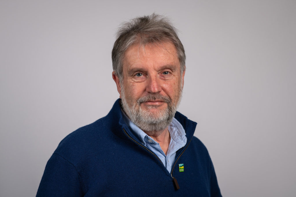 Head and shoulders of older bearded man smiling, in blue shirt and casual top