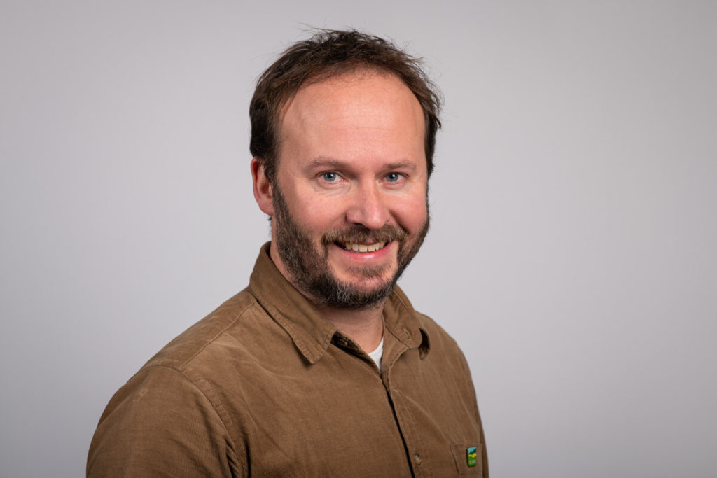 Man with trimmed beard and brown shirt smiling at camera