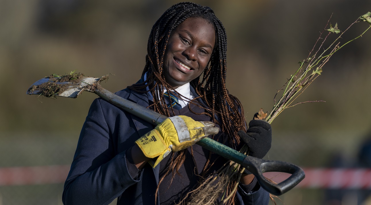 Teenage girl carrying spade and bare root plant smiling at camera.