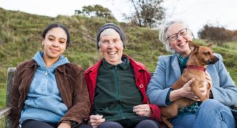 Three women of different generations sitting on a bench in the countryside smiling
