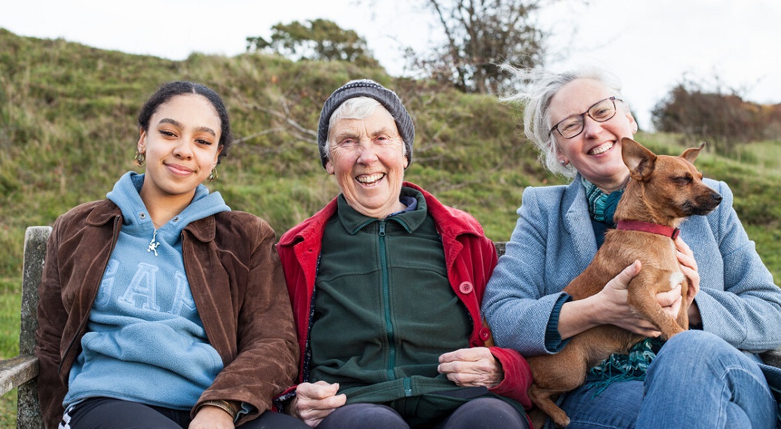 Three women of different generations sitting on a bench in the countryside smiling