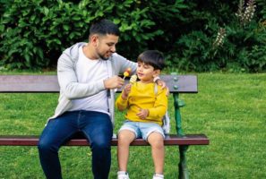 Young father and son sitting on bench in park sharing ice lollies