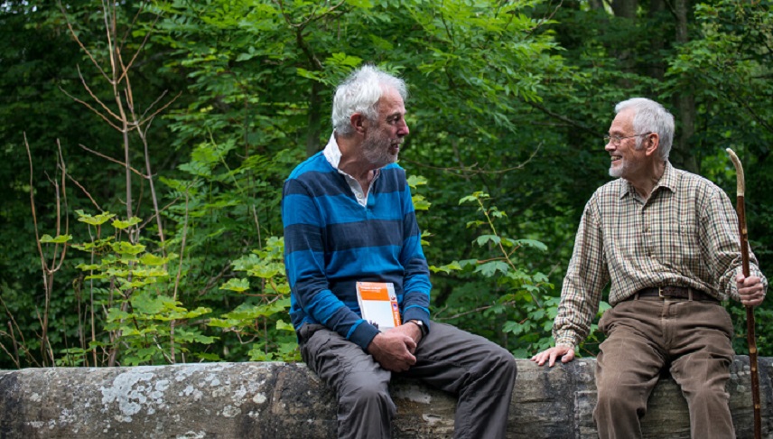 Two older men sit on a wall in front of trees holding a map and walking stick