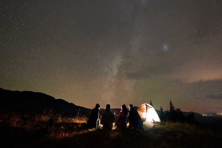 Small group of silhouetted people gazing at the night sky sitting outside a lit tent.