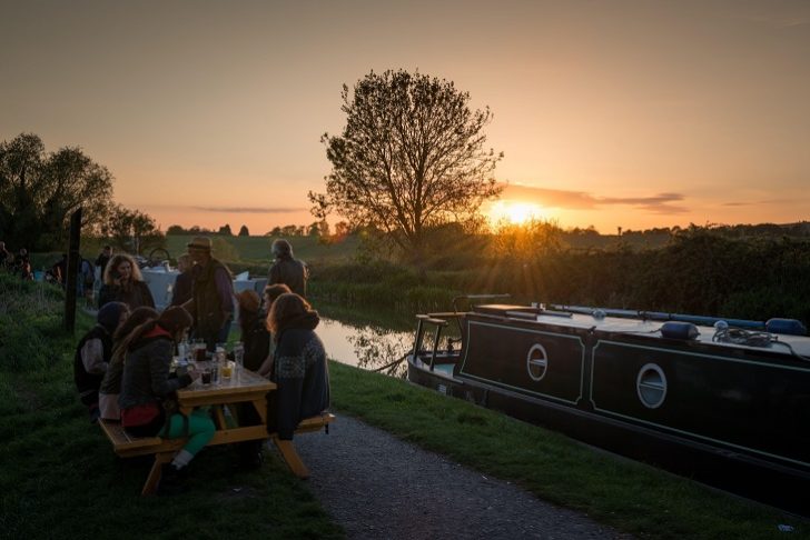 People sitting drinking by a narrow boat on a canal in the countryside at sunset.