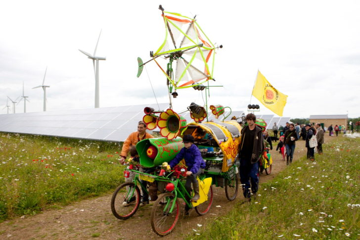 A community push a brightly decorated trailer along with solar panels and wind turbines behind