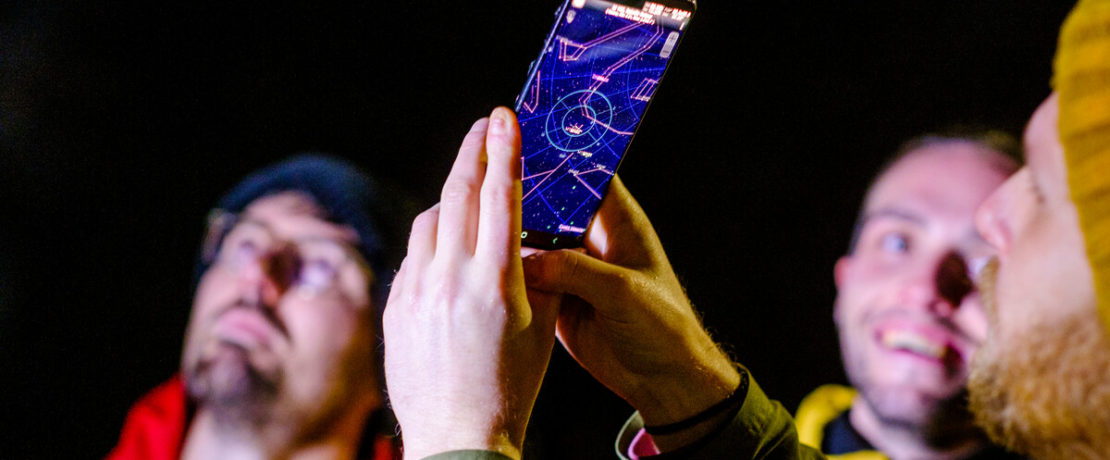 Man holding phone up to sky to view the star constellations with men in background