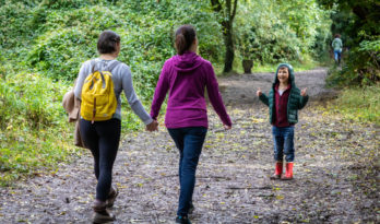 Two women with back to camera walking along wooded path kid with thumbs up