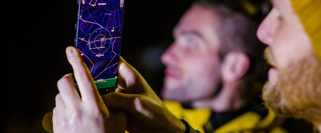 Man holding phone up to sky to view the star constellations with men in background