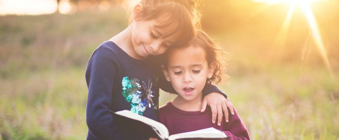 Two young children reading a book in a field with the sun shining brightly in the background