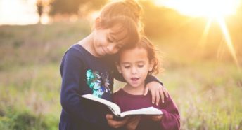 Two young children reading a book in a field with the sun shining brightly in the background