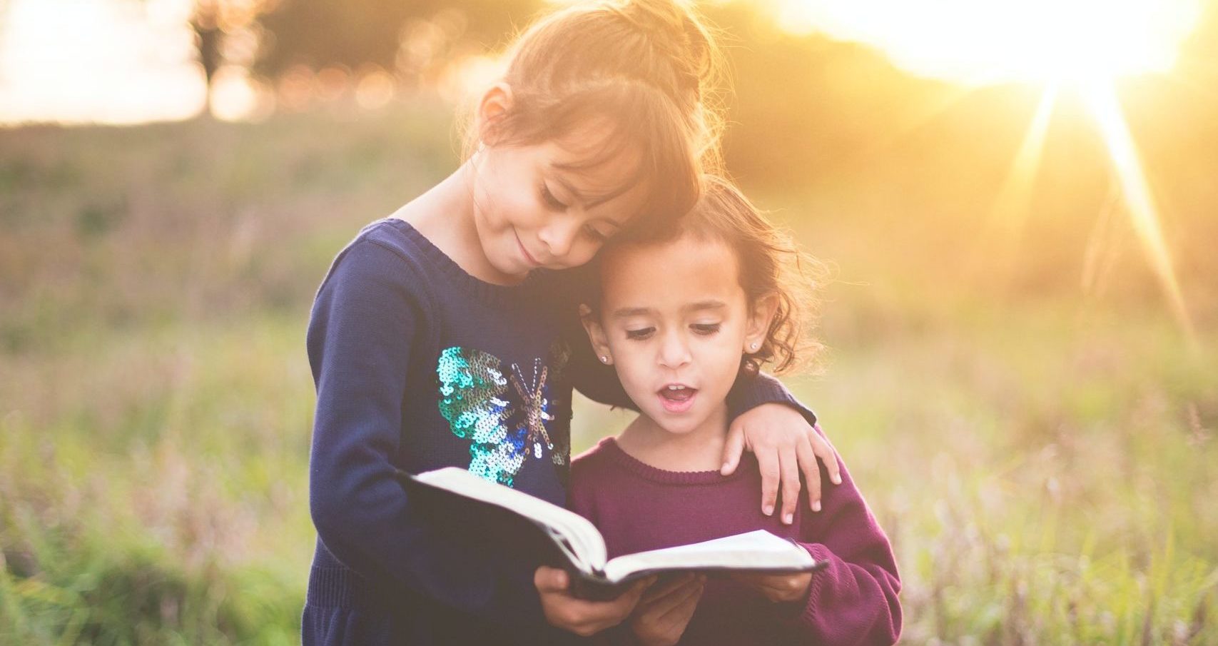 Two young children reading a book in a field with the sun shining brightly in the background