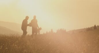 Two people walking across a sun drenched field at harvest