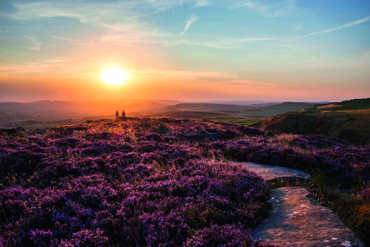 Absolutely stunning sunset landscape image with unidentified couple looking from Higger Tor in Peak District across to Hope Vally in late Summer with heather in full bloom