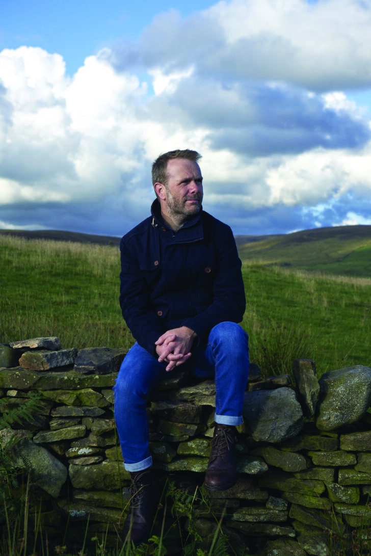 Photograph of Andrew Michael Hurley sitting on a stonewall in the countryside
